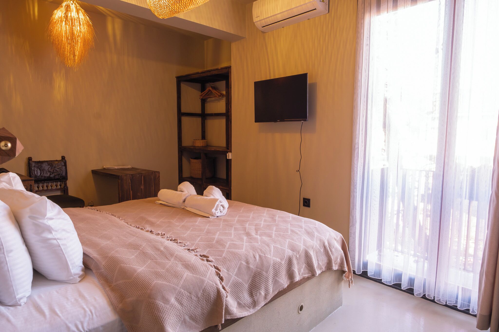 A hotel room with a large bed, a TV mounted on the wall, a shelf with baskets, and a window with sheer curtains.