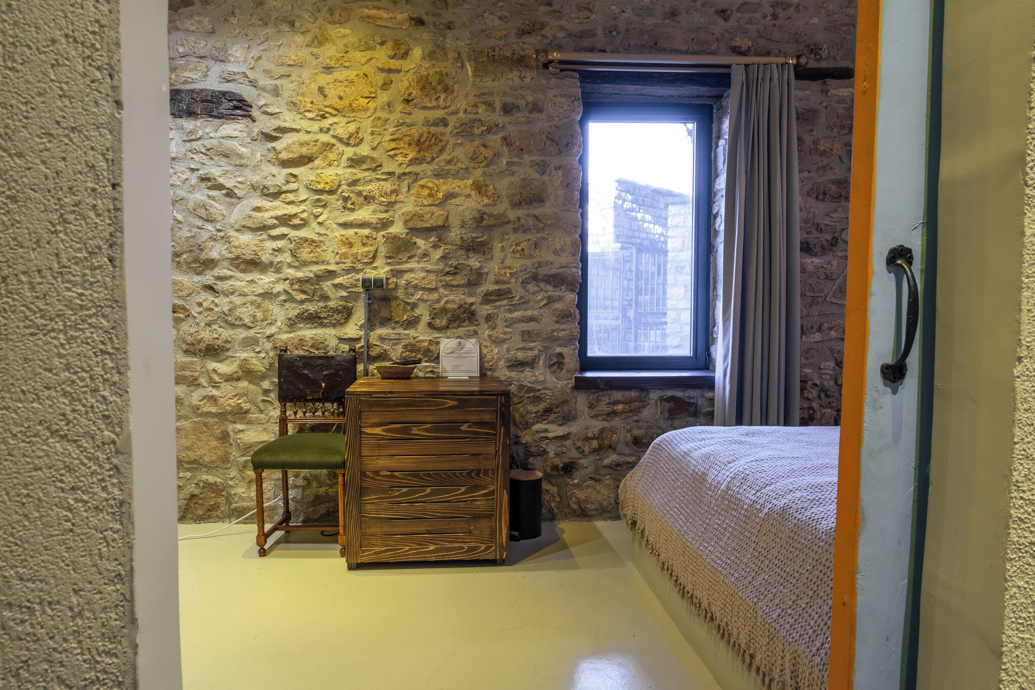 A room with a stone wall, a wooden dresser with a green chair, a bed with a white blanket, and a window with curtains.