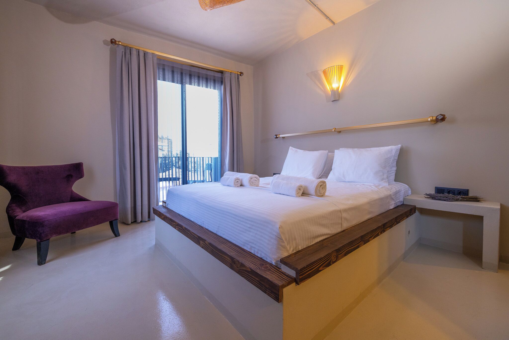 A hotel room with a bed, a purple chair, a bedside table, and a view of the city through the window.