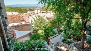 A courtyard with stone walls, a grassy area, and a view of a town with terracotta roofs. - Belle Maison Charmante Typiquement Provençale (Meyrargues)