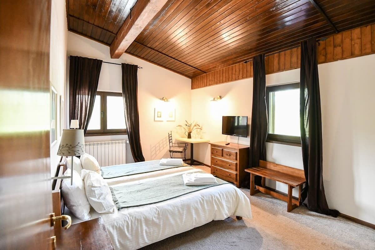 A bedroom with a wooden ceiling, a bed with white bedding, a desk with a chair, a TV, and a window with curtains.
