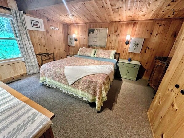 Un dormitorio acogedor con una cama grande, paredes de madera, una mesita de noche pequeña y una ventana con cortinas.