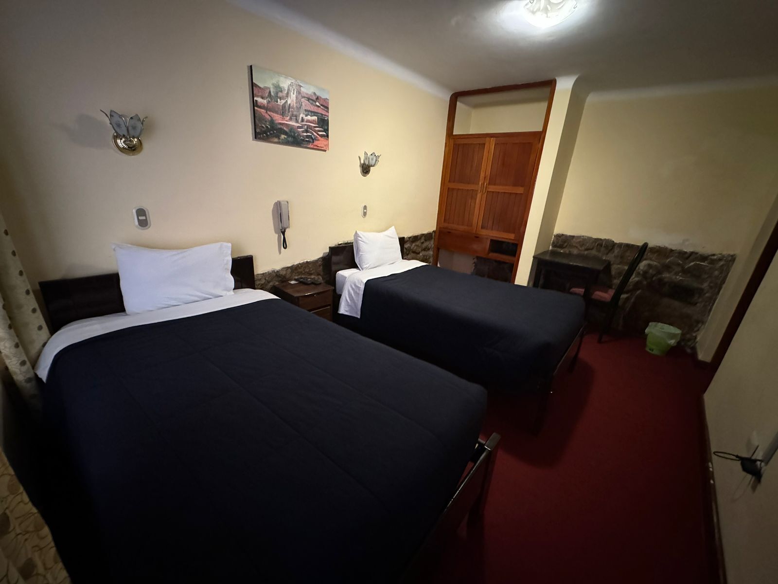 A hotel room with two beds, a wooden wardrobe, a telephone, and a painting on the wall.