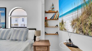 A bedroom with a bed, a bedside table, a lamp, a bookshelf, and a large beach painting on the wall. - Destin/Miramar Beach Townhouse, 3-Min Walk to Sand, Sleeps 6, Pet Friendly (Miramar Beach)