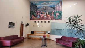 A hotel lobby with a reception desk, two red sofas, a small table with a vase, a large mural depicting a cultural scene, and a sign for Hotel Plaza Pachuca. - Línea Divisoria (Zapotlan de Juarez)