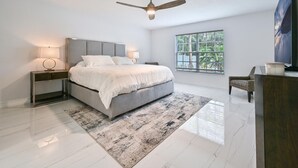 A modern bedroom with a bed, nightstands, a ceiling fan, a window with a view of greenery, and a television on a wooden stand. - Spacious+cozy house, near the beach, heated pool. (Naples)