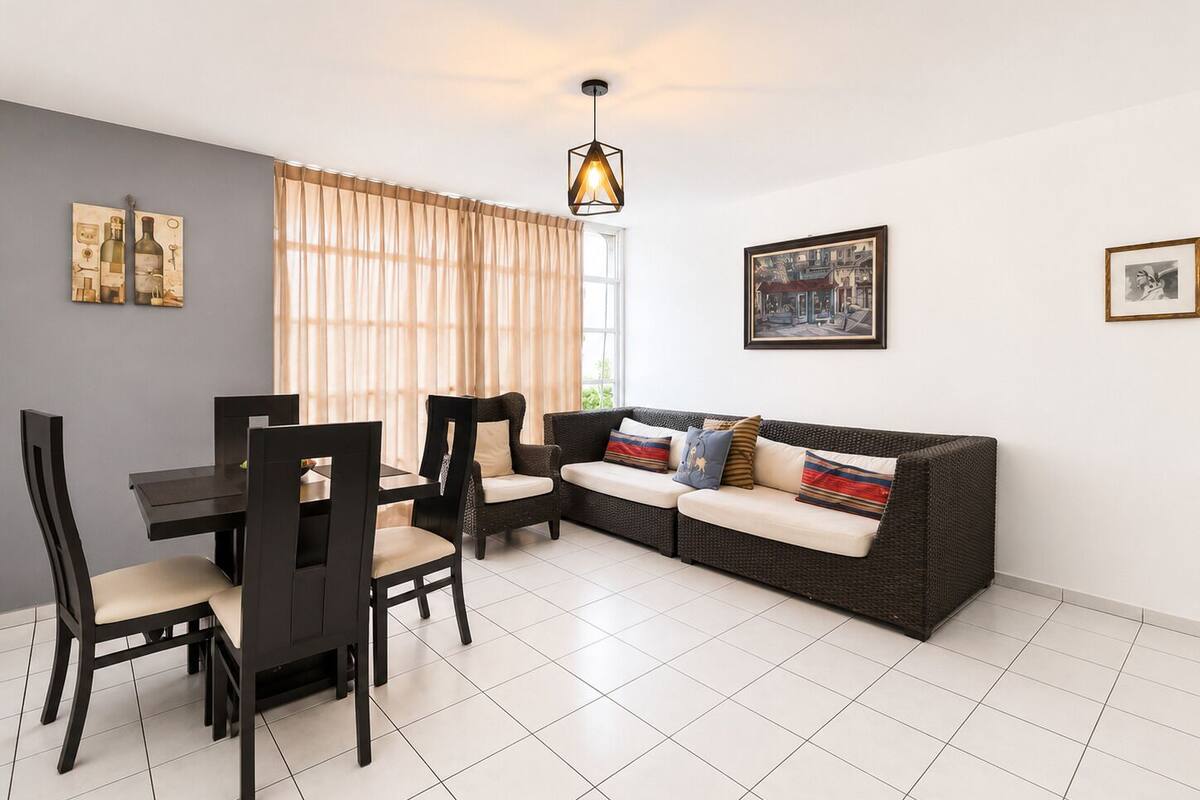 A dining area with a table and chairs, a sofa with cushions, a hanging light fixture, and two framed pictures on the walls.