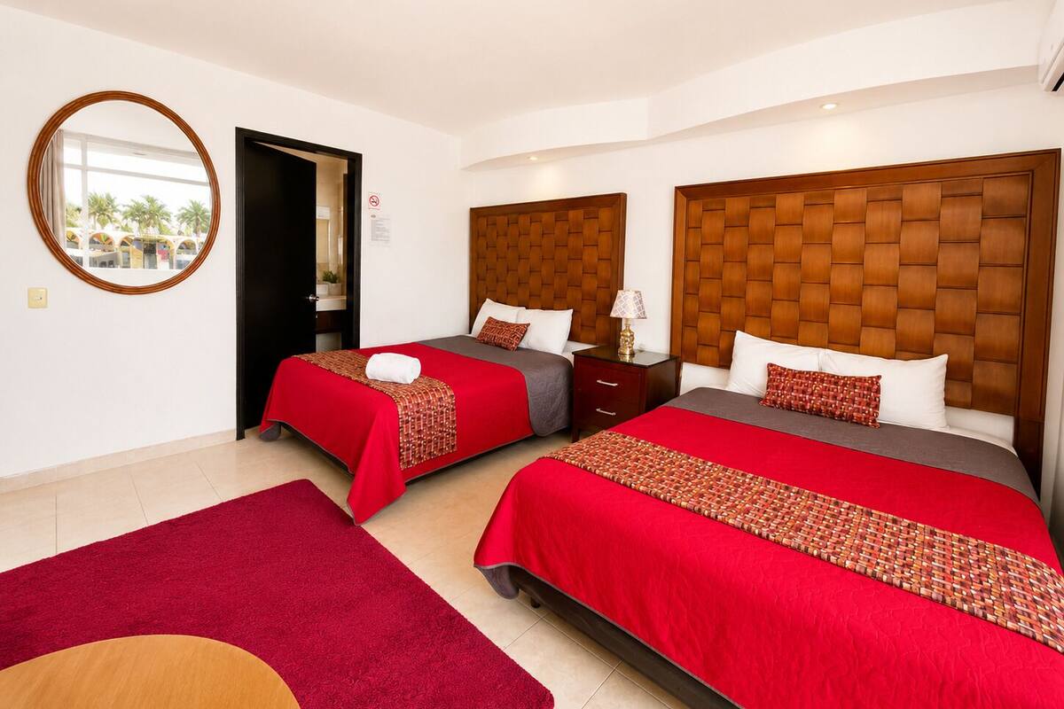 A hotel room with two beds, a round mirror, a red carpet, and a view of palm trees outside.