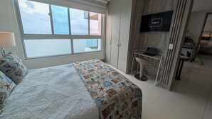 Un dormitorio con un ventanal grande, una cama con colcha, un escritorio con computadora portátil y un televisor montado en la pared.