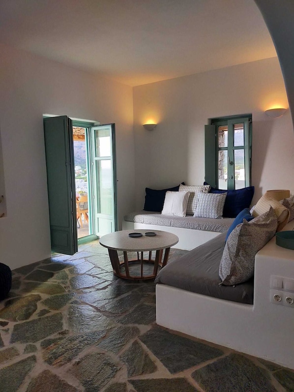 A cozy living room with a stone floor, a round coffee table, a sofa with patterned cushions, and a view through a door. - Amorgos Elegant Houses, Villa 3 (Amorgos)