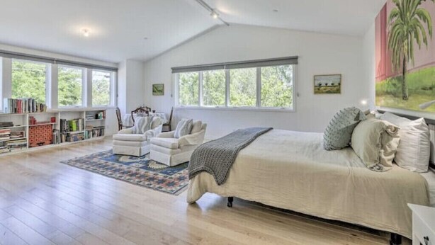 Un dormitorio amplio con una cama grande, un rincón de descanso con una silla y una mesita, una estantería y ventanas grandes que ofrecen vistas al verdor.