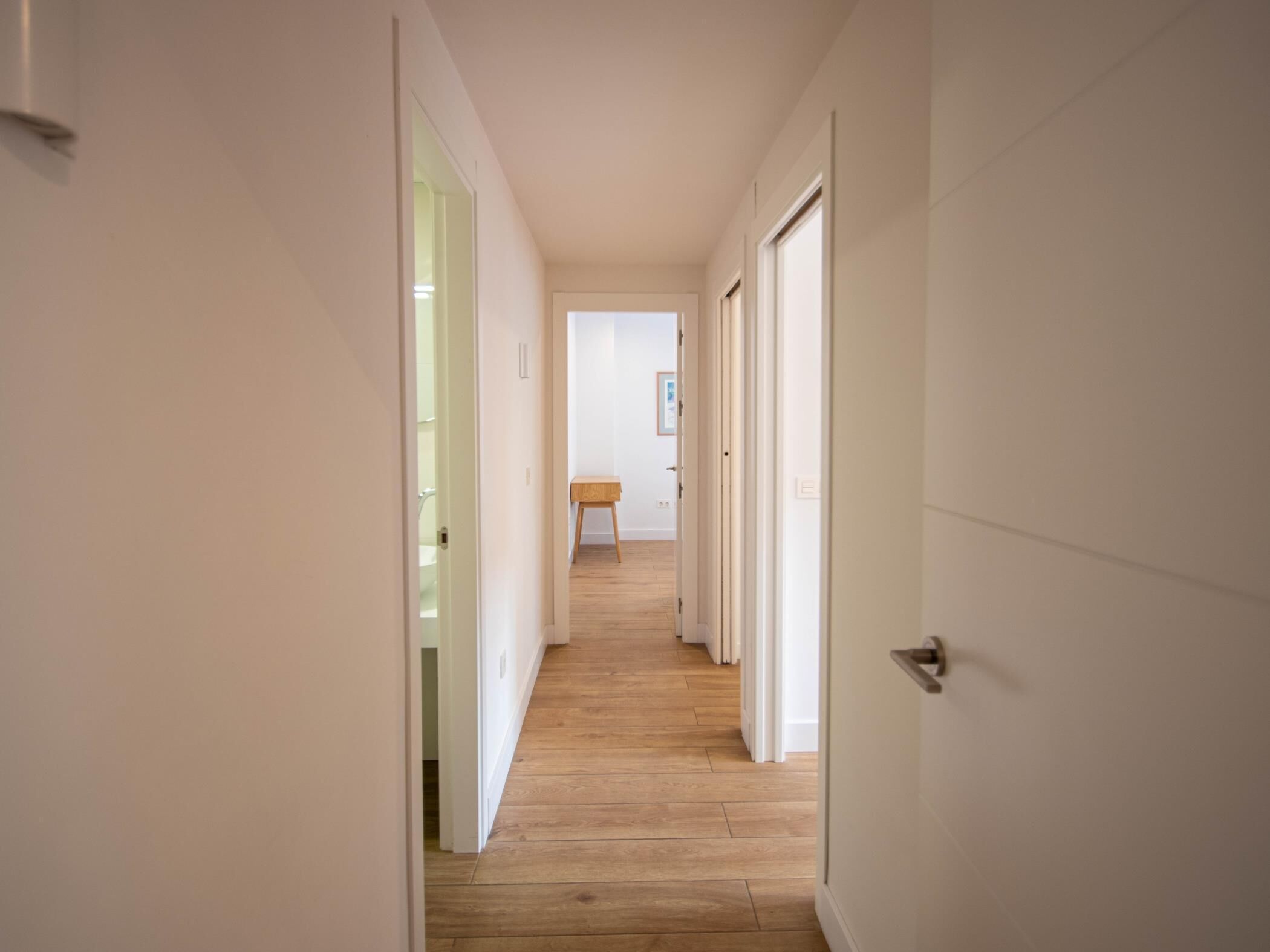 A narrow hallway with wooden flooring and white walls, leading to a room with a wooden table and a framed picture on the wall.