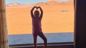 A room with a view of a desert landscape, a window with curtains, and a person making a heart shape with their hands.