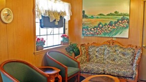 A cozy room with a wooden wall, a floral sofa, two green armchairs, a wooden coffee table, a clock, and a window with a view of a field. - American Dream (Elk City)