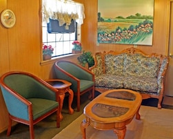 A cozy room with a wooden wall, a floral sofa, two green armchairs, a wooden coffee table, a clock, and a window with a view of a field.