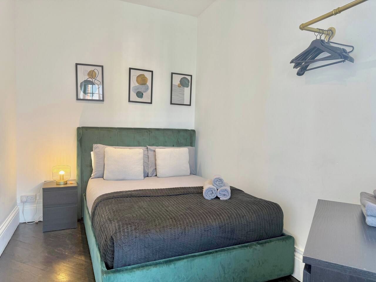 Un lit bien fait, avec une tête de lit verte, une table de chevet avec une lampe et un porte-manteau fixé au mur.