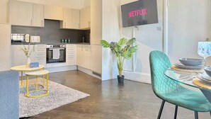 A modern kitchen with white cabinets, a dining area with a glass table and green chairs, and a TV mounted on the wall displaying 'NETFLIX'. - Sleek 1 Bedroom flat in Shoreditch, Old Street (London)