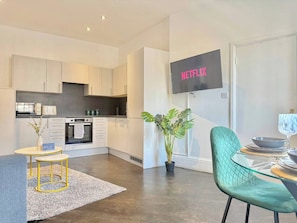 A modern kitchen with white cabinets, a dining area with a glass table and green chairs, and a TV mounted on the wall displaying 'NETFLIX'. - Sleek 1 Bedroom flat in Shoreditch, Old Street (London)