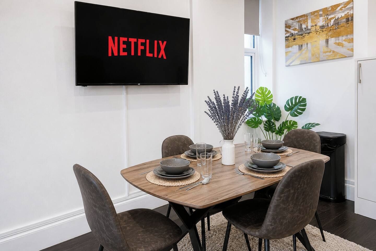 A modern dining area with a wooden table set for four, a flat-screen TV displaying 'NETFLIX,' and a decorative plant.