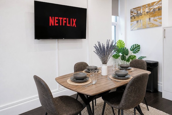 A modern dining area with a wooden table set for four, a flat-screen TV displaying 'NETFLIX,' and a decorative plant.
