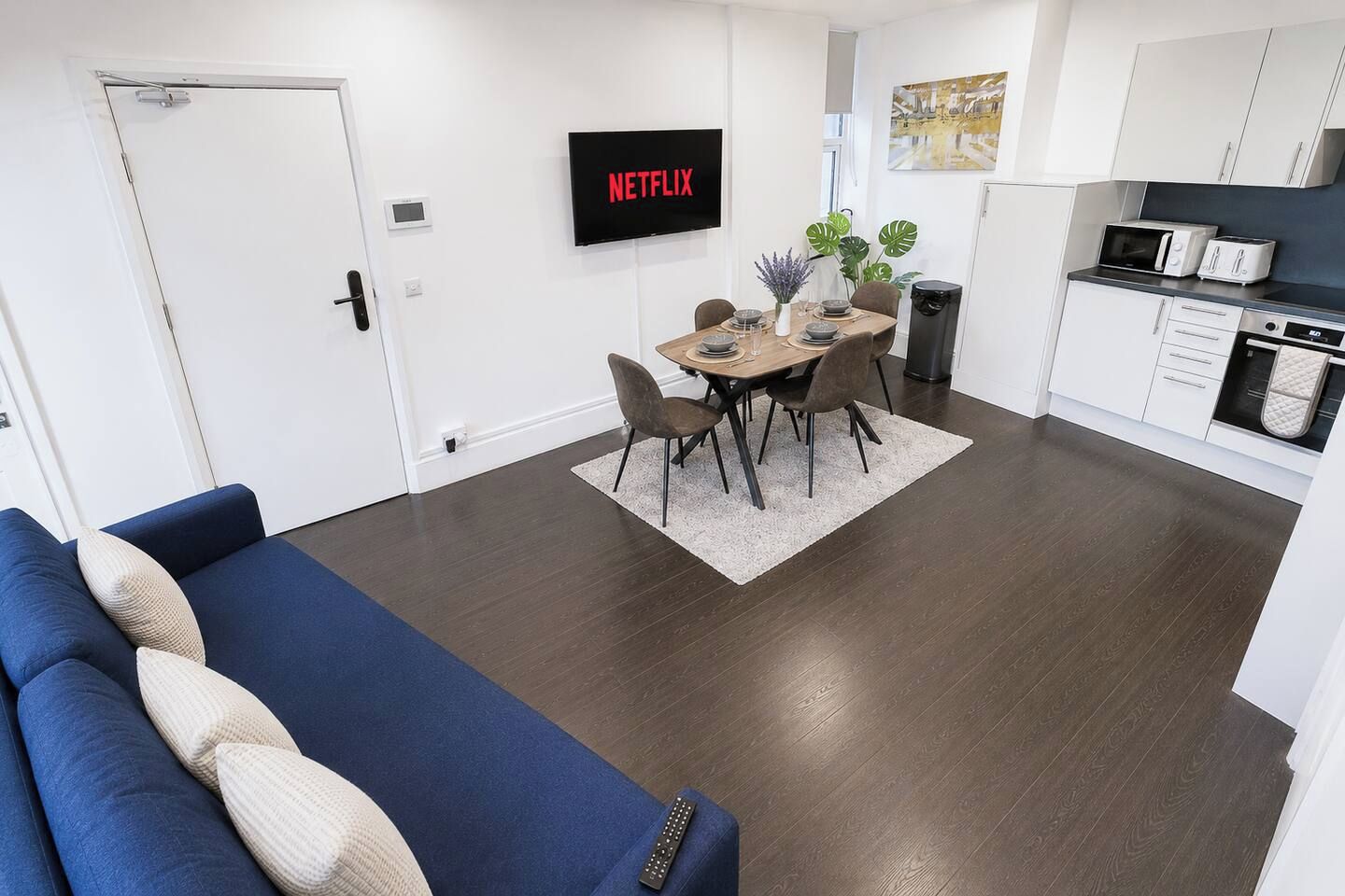 A modern living room with a blue sofa, a dining area with a table and chairs, a kitchen with white cabinets and appliances, and a TV mounted on the wall displaying 'NETFLIX'.