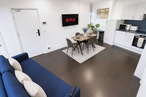 A modern living room with a blue sofa, a dining area with a table and chairs, a kitchen with white cabinets and appliances, and a TV mounted on the wall displaying 'NETFLIX'.
