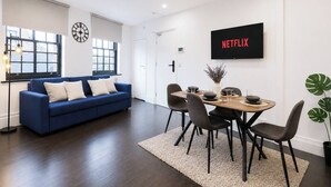 A modern living room with a blue sofa, a dining table with chairs, a wall-mounted TV, and a large window with blinds.