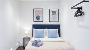 A neatly made bed with a blue headboard, two pillows, and rolled towels. There is a bedside table with a lamp and a framed picture on the wall.