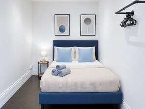 A neatly made bed with a blue headboard, two pillows, and rolled towels. There is a bedside table with a lamp and a framed picture on the wall.