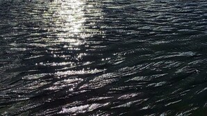 Uma praia com uma grande formação rochosa, ondas e reflexos de luz solar na água.