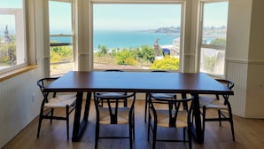Uma sala de jantar com uma mesa grande e quatro cadeiras, vista para o mar e vegetação, e uma estátua de um pelicano.
