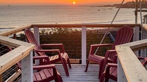 Um deck de madeira com cadeiras vermelhas e guarda-corpo, com vista para o oceano ao pôr do sol.