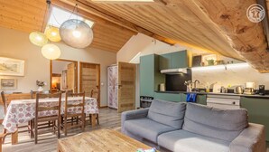 A cozy living room with a sofa, wooden dining table, and a kitchen area with a modern stove. - Residence Mont- Blanc Lodge - LA Fleche (HAUTELUCE)