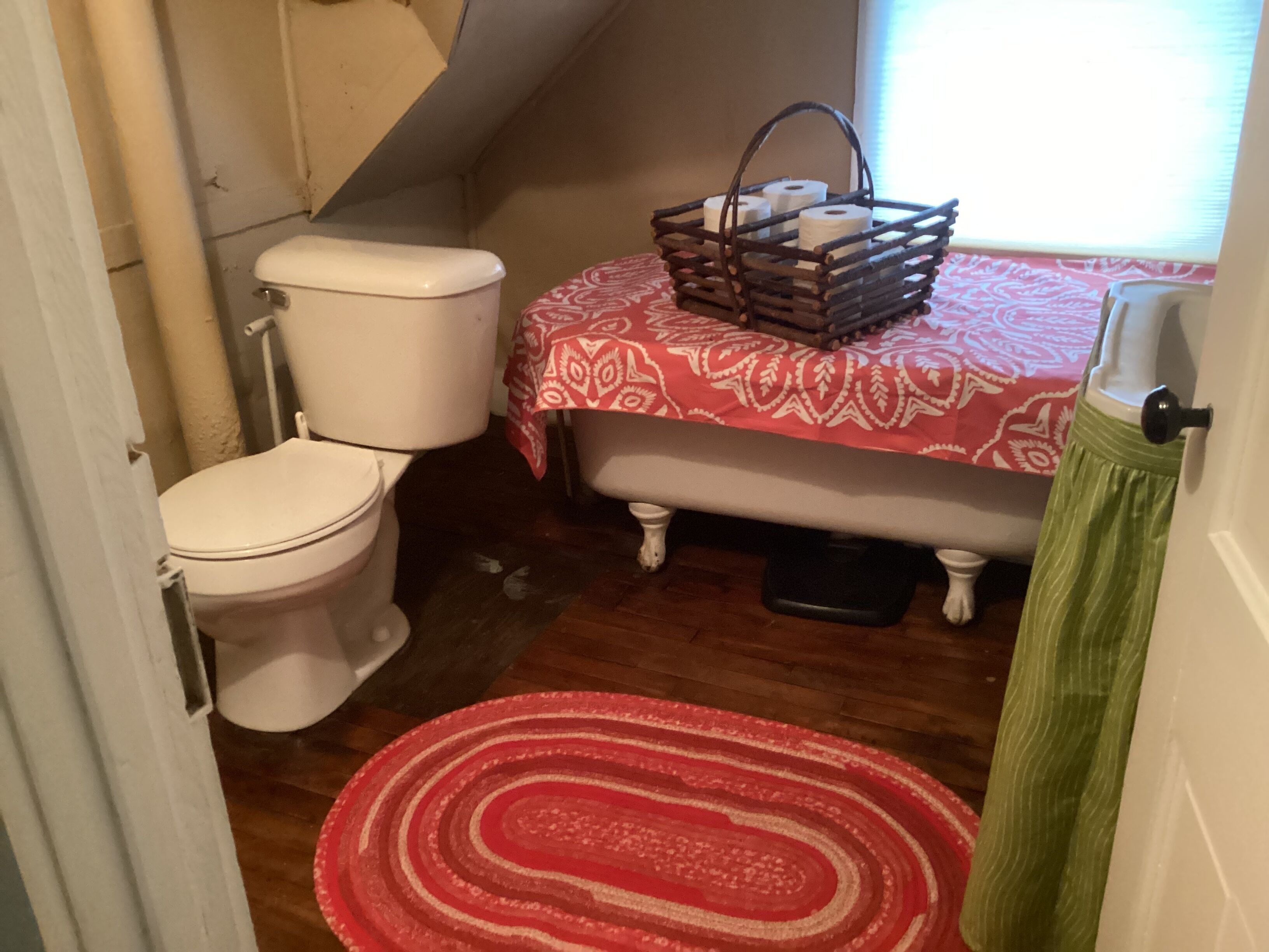A small bathroom with a toilet, a wooden bench with a basket, and a red patterned rug.