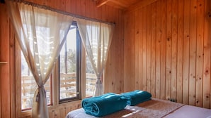 Une pièce de cabane en bois comprenant un lit, une fenêtre avec des rideaux et donnant sur une terrasse en bois à l’extérieur.