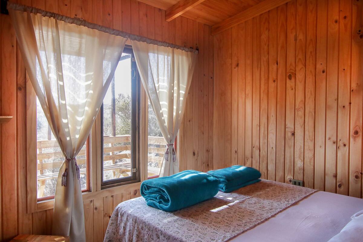 Een houten kamer met een bed, een raam met gordijnen en uitzicht op een houten terras.