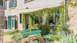 Ein Innenhof mit einem runden Tisch und Stühlen, umgeben von Grünpflanzen und einem Gebäude mit mit Efeu bewachsenen Fenstern.
