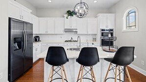Una cocina moderna con armarios blancos, un refrigerador de acero inoxidable, una isla central con sillas negras y una lámpara colgante.