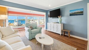 A living room with a view of the sea, a white sofa, a light blue armchair, a white coffee table, and a cabinet with a flat-screen TV. - Amazing Marina Views | Baypoint Resort | Golf Course (Panama City Beach)
