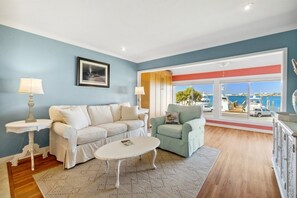 A living room with a white sofa, a green armchair, and a white coffee table. There is a large window with a view of the sea and boats.