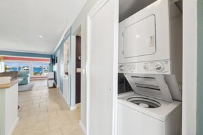 A laundry room with a front-loading washing machine, a tiled floor, and a view of the sea through an open door.
