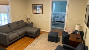 A living room with a gray sectional sofa, a small ottoman, a coffee table, and a side table with a lamp. There is a framed picture on the wall and a window with blinds.