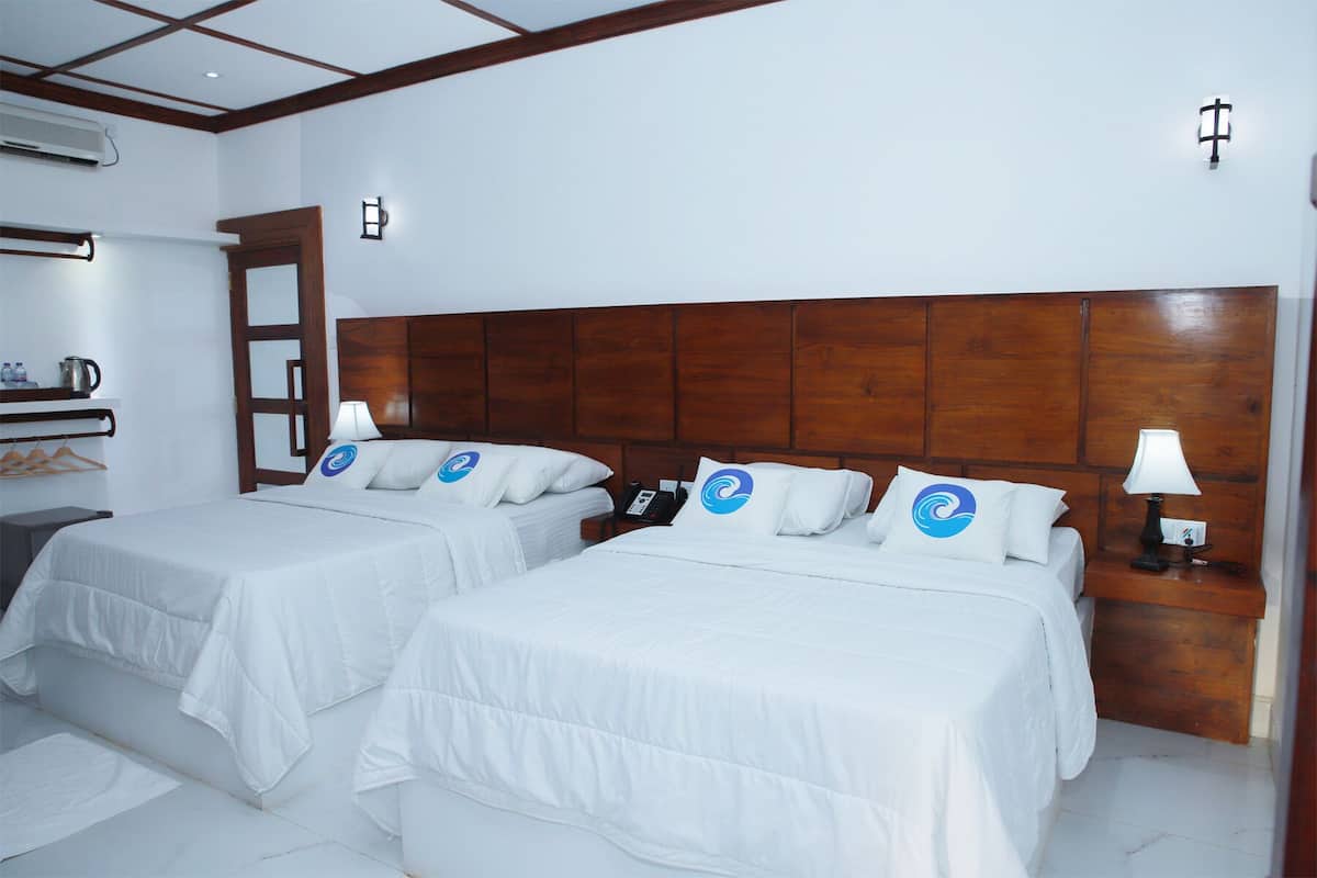 A hotel room with two beds, wooden headboards, white bedding, a bedside table with a lamp, and a wall-mounted air conditioning unit.