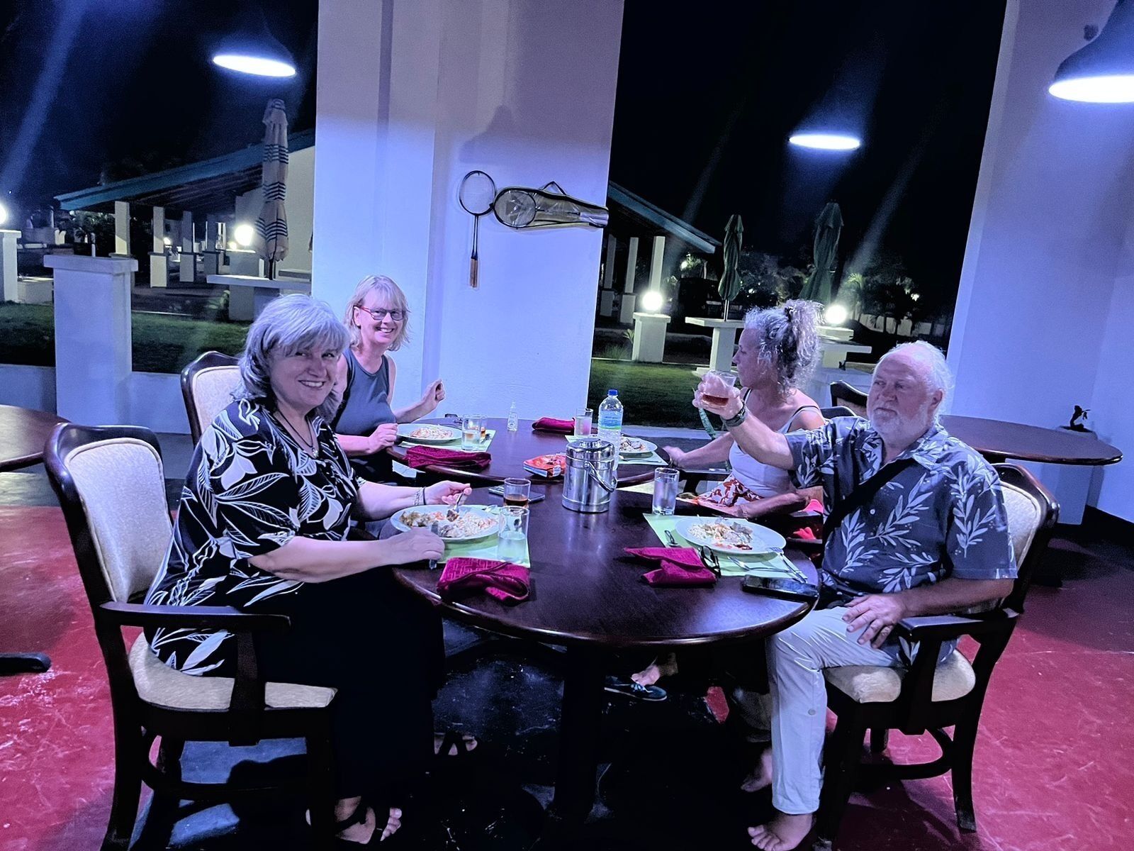 Un grupo de personas cenando en una mesa al aire libre con sillas, una lámpara de mesa y una botella de agua sobre la mesa.