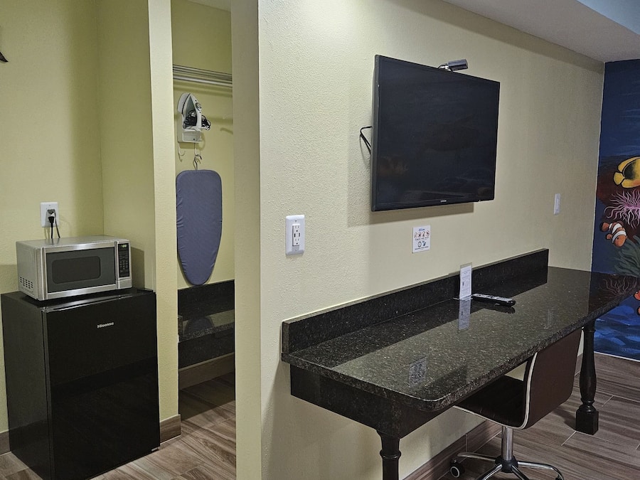 A room with a flat-screen TV mounted on the wall, a small kitchenette with a microwave, and a desk with a chair.