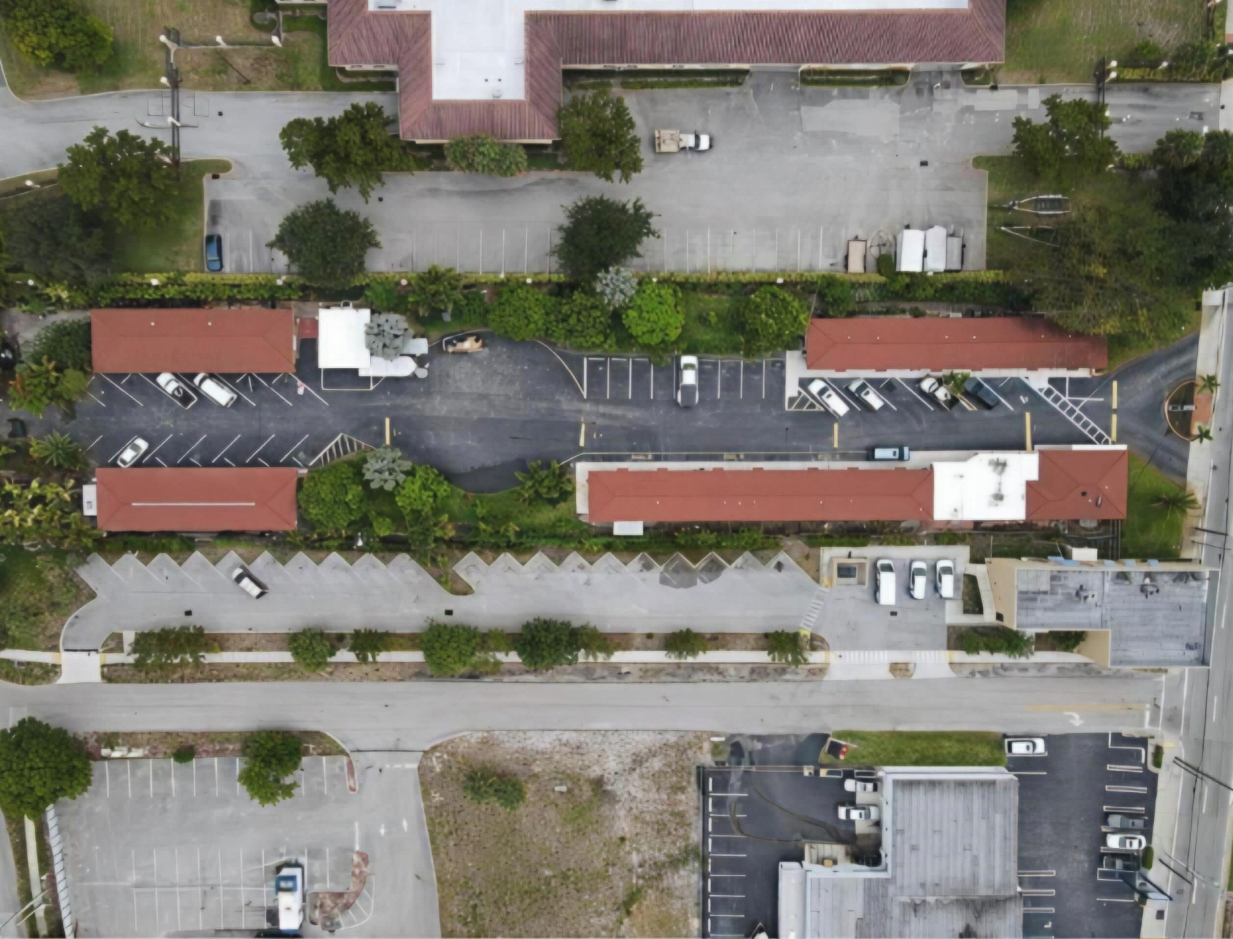 An aerial view of a parking lot with multiple rows of parking spaces, a central pedestrian pathway, and surrounding greenery.