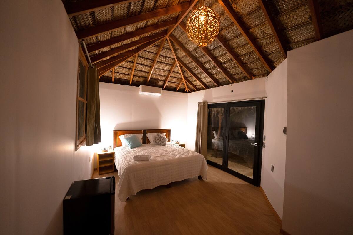 A bedroom with a wooden ceiling, a bed with a white bedspread, a nightstand, and a sliding glass door leading to a balcony.