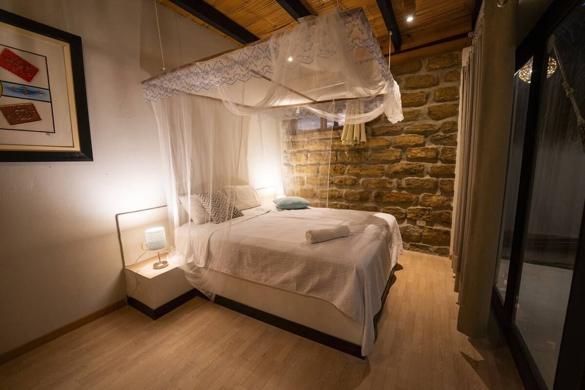 A bedroom with a four-poster bed, a nightstand with a lamp, and a stone wall.