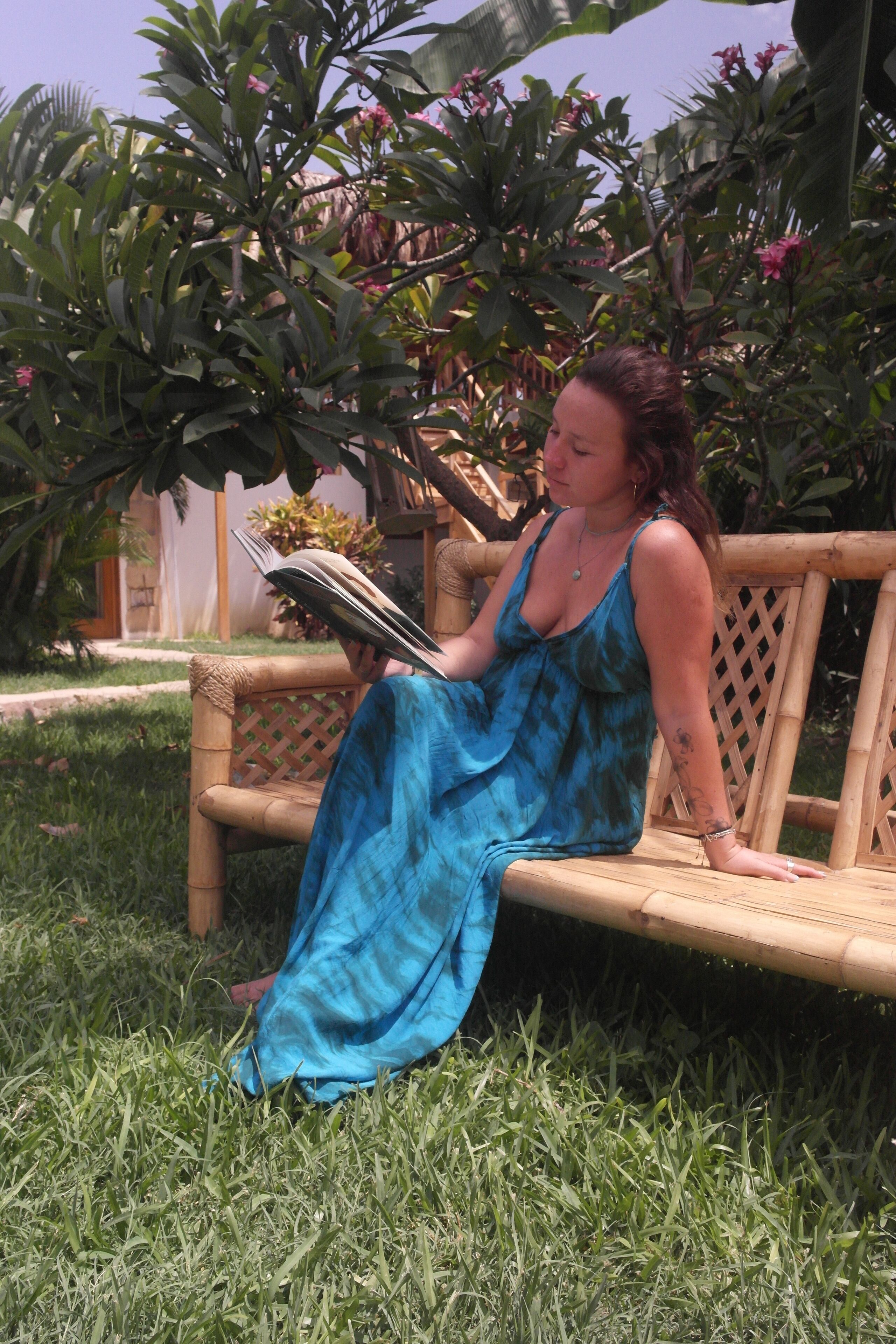A person sitting on a wooden bench in a garden area with greenery and a building in the background.