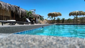 A swimming pool with lounge chairs and thatched umbrellas, surrounded by a wooden fence and palm trees. - Panorama Garden Inn (Máncora)